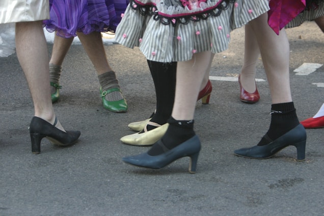 A group of people are standing closely together, wearing colorful and varied footwear, including high heels and socks. Skirts with different patterns and textures are visible, adding a playful and festive atmosphere.