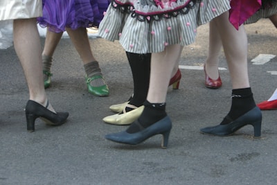 A group of people are standing closely together, wearing colorful and varied footwear, including high heels and socks. Skirts with different patterns and textures are visible, adding a playful and festive atmosphere.