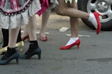 A group of people wear various brightly colored high-heeled shoes while walking on a paved street. The legs are clad in socks or tights, and skirts with decorative trim are visible. A car tire is seen in the background.
