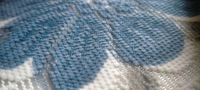 A close-up view of a fabric pattern featuring woven threads in shades of blue and white. The texture is visible, with intricate stitching creating a soft, tactile appearance. The fibers appear dense and tightly knit.