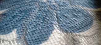 A close-up view of a fabric pattern featuring woven threads in shades of blue and white. The texture is visible, with intricate stitching creating a soft, tactile appearance. The fibers appear dense and tightly knit.