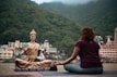 A person is sitting cross-legged on a yoga mat, meditating in front of a large statue of a serene figure. The background features lush green mountains and several buildings, indicating a tranquil setting by the river.