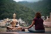 A person is sitting cross-legged on a yoga mat, meditating in front of a large statue of a serene figure. The background features lush green mountains and several buildings, indicating a tranquil setting by the river.