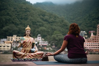 A person is sitting cross-legged on a yoga mat, meditating in front of a large statue of a serene figure. The background features lush green mountains and several buildings, indicating a tranquil setting by the river.