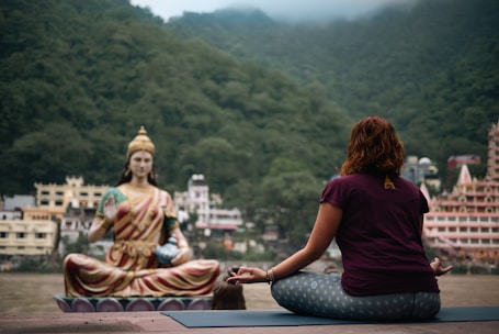 A person is sitting cross-legged on a yoga mat, meditating in front of a large statue of a serene figure. The background features lush green mountains and several buildings, indicating a tranquil setting by the river.