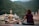 A person is sitting cross-legged on a yoga mat, meditating in front of a large statue of a serene figure. The background features lush green mountains and several buildings, indicating a tranquil setting by the river.