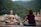 A person is sitting cross-legged on a yoga mat, meditating in front of a large statue of a serene figure. The background features lush green mountains and several buildings, indicating a tranquil setting by the river.