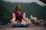 A woman sits cross-legged on a yoga mat in an outdoor setting, surrounded by green mountains and buildings in the background. She is dressed in a maroon top and patterned leggings, with her eyes closed and hands resting on her knees in a meditative pose.