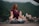 A woman sits cross-legged on a yoga mat in an outdoor setting, surrounded by green mountains and buildings in the background. She is dressed in a maroon top and patterned leggings, with her eyes closed and hands resting on her knees in a meditative pose.
