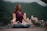 A woman sits cross-legged on a yoga mat in an outdoor setting, surrounded by green mountains and buildings in the background. She is dressed in a maroon top and patterned leggings, with her eyes closed and hands resting on her knees in a meditative pose.