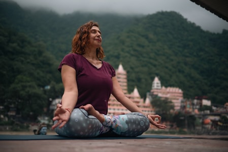 A woman sits cross-legged on a yoga mat in an outdoor setting, surrounded by green mountains and buildings in the background. She is dressed in a maroon top and patterned leggings, with her eyes closed and hands resting on her knees in a meditative pose.
