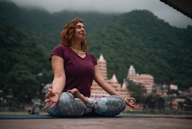 A woman sits cross-legged on a yoga mat in an outdoor setting, surrounded by green mountains and buildings in the background. She is dressed in a maroon top and patterned leggings, with her eyes closed and hands resting on her knees in a meditative pose.