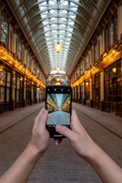 A person is holding a smartphone in their hands, capturing an image of a long, covered arcade with a glass roof and intricate architectural detailing. The view through the phone aligns perfectly with the real scene, creating a sense of depth and perspective. Warm lighting illuminates the arcade, casting a welcoming glow.