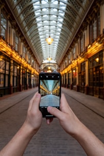 A person is holding a smartphone in their hands, capturing an image of a long, covered arcade with a glass roof and intricate architectural detailing. The view through the phone aligns perfectly with the real scene, creating a sense of depth and perspective. Warm lighting illuminates the arcade, casting a welcoming glow.