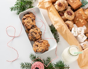 A variety of cookies and sweets are artfully arranged, including chocolate chip cookies in a white tray, pecan-topped cookies, and nougat pieces, alongside a milk bottle with a green-striped straw. Decorative elements include sprigs of evergreen and red-and-white twine, creating a festive atmosphere.