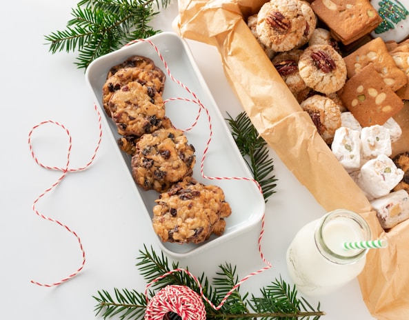 A variety of cookies and sweets are artfully arranged, including chocolate chip cookies in a white tray, pecan-topped cookies, and nougat pieces, alongside a milk bottle with a green-striped straw. Decorative elements include sprigs of evergreen and red-and-white twine, creating a festive atmosphere.