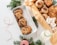 A variety of cookies and sweets are artfully arranged, including chocolate chip cookies in a white tray, pecan-topped cookies, and nougat pieces, alongside a milk bottle with a green-striped straw. Decorative elements include sprigs of evergreen and red-and-white twine, creating a festive atmosphere.