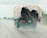 A truck carrying a load of logs covered with a tarp drives down a wet highway, creating a mist of water behind it. The road is flanked by greenery and other vehicles can be seen in the distance.