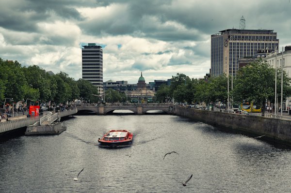 A boat traveling down a river next to tall buildings — Dublin, Ireland