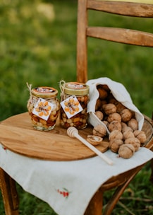 Two jars filled with mixed nuts and honey are placed on a wooden chair with a wooden plate underneath. A honey dipper rests beside the jars. A white linen bag, partially spilling walnuts, is placed next to the jars.