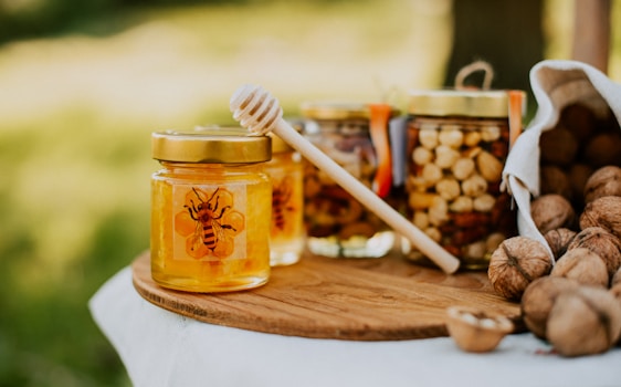 A jar of honey with a bee design on the label sits on a wooden board. Next to it, a wooden honey dipper leans against the jar. In the background, there are jars filled with nuts and an overturned cloth bag spilling out walnuts. The scene is set outdoors with a blurred green background.