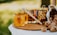 A jar of honey with a bee design on the label sits on a wooden board. Next to it, a wooden honey dipper leans against the jar. In the background, there are jars filled with nuts and an overturned cloth bag spilling out walnuts. The scene is set outdoors with a blurred green background.
