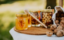 A jar of honey with a bee design on the label sits on a wooden board. Next to it, a wooden honey dipper leans against the jar. In the background, there are jars filled with nuts and an overturned cloth bag spilling out walnuts. The scene is set outdoors with a blurred green background.