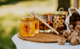 A jar of honey with a bee design on the label sits on a wooden board. Next to it, a wooden honey dipper leans against the jar. In the background, there are jars filled with nuts and an overturned cloth bag spilling out walnuts. The scene is set outdoors with a blurred green background.