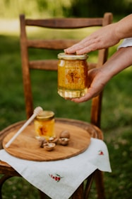 A glass jar filled with golden honey is being held by two hands over a wooden chair placed outdoors. On the seat of the chair, there is a wooden tray with another jar of honey, walnuts, and a honey dipper resting nearby. A white cloth with small red berry embroidery partially covers the chair seat.