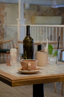 A cozy cafe setting featuring a table with a cup of coffee in the foreground. Besides the coffee, there are several glass and ceramic items, including a dark green bottle, a pinkish mug, a clear glass, and a metal container holding straws. The ambient background is a blend of rustic and modern elements, with wooden chairs and a lightly textured wall.