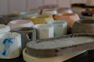 A collection of various ceramic cups and bowls in different shapes and colors arranged closely together on a dark surface. The ceramics appear handcrafted with a rustic, unfinished texture. Hues range from soft whites and grays to blues, yellows, and muted oranges.