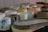 A collection of various ceramic cups and bowls in different shapes and colors arranged closely together on a dark surface. The ceramics appear handcrafted with a rustic, unfinished texture. Hues range from soft whites and grays to blues, yellows, and muted oranges.