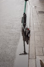 A green electric scooter is parked next to a curb on a wet sidewalk with a puddle reflecting the scooter. The pavement appears damp, suggesting recent rainfall, and there are rain droplets visible on the water's surface.