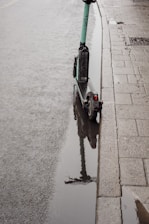 A green electric scooter is parked next to a curb on a wet sidewalk with a puddle reflecting the scooter. The pavement appears damp, suggesting recent rainfall, and there are rain droplets visible on the water's surface.