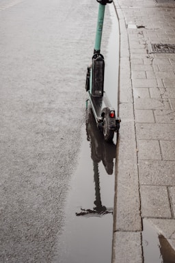 A green electric scooter is parked next to a curb on a wet sidewalk with a puddle reflecting the scooter. The pavement appears damp, suggesting recent rainfall, and there are rain droplets visible on the water's surface.