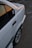 A close-up view of a white car parked on a textured pavement. The car's rear side is visible with a sticker on the window and a hint of sunset reflecting off the surface.
