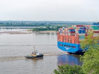 A large cargo ship laden with multicolored shipping containers is being guided along a river by a small tugboat. The river is calm and wide, while green trees line the banks in the background, providing a lush setting. The sky is overcast, casting a serene and somewhat muted light over the scene.