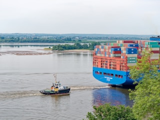 A large cargo ship laden with multicolored shipping containers is being guided along a river by a small tugboat. The river is calm and wide, while green trees line the banks in the background, providing a lush setting. The sky is overcast, casting a serene and somewhat muted light over the scene.