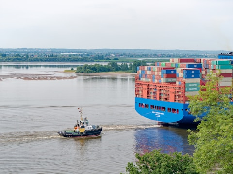 A large cargo ship laden with multicolored shipping containers is being guided along a river by a small tugboat. The river is calm and wide, while green trees line the banks in the background, providing a lush setting. The sky is overcast, casting a serene and somewhat muted light over the scene.