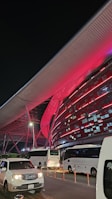 A modern architectural structure illuminated with vibrant red lights, featuring sleek metallic elements and a large overhanging roof. Several white buses and a white car are parked in front of the building, indicating possible transportation services. The scene is set at night, adding to the dramatic lighting effect.