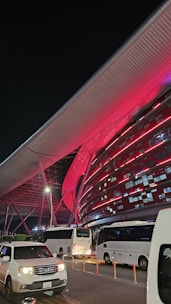 A modern architectural structure illuminated with vibrant red lights, featuring sleek metallic elements and a large overhanging roof. Several white buses and a white car are parked in front of the building, indicating possible transportation services. The scene is set at night, adding to the dramatic lighting effect.