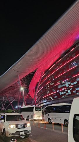 A modern architectural structure illuminated with vibrant red lights, featuring sleek metallic elements and a large overhanging roof. Several white buses and a white car are parked in front of the building, indicating possible transportation services. The scene is set at night, adding to the dramatic lighting effect.