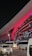 A modern architectural structure illuminated with vibrant red lights, featuring sleek metallic elements and a large overhanging roof. Several white buses and a white car are parked in front of the building, indicating possible transportation services. The scene is set at night, adding to the dramatic lighting effect.