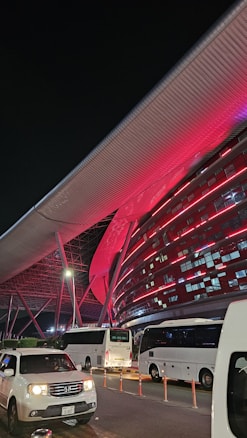 A modern architectural structure illuminated with vibrant red lights, featuring sleek metallic elements and a large overhanging roof. Several white buses and a white car are parked in front of the building, indicating possible transportation services. The scene is set at night, adding to the dramatic lighting effect.