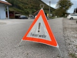 A triangular warning sign with an exclamation mark and the word 'Feuerwehr' printed on it stands on a paved road. Surrounding the sign is an outdoor setting with a road, vehicles, green hills, trees, and houses in the background.
