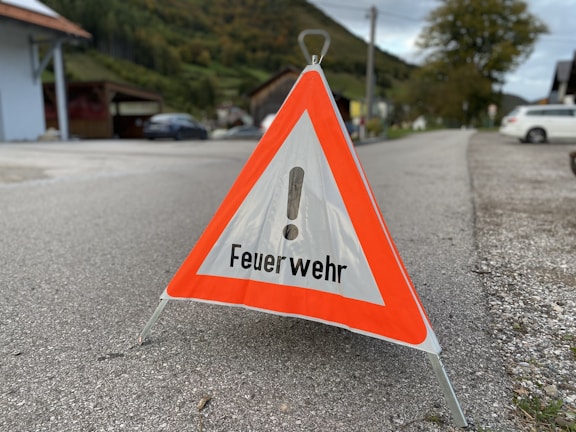 A triangular warning sign with an exclamation mark and the word 'Feuerwehr' printed on it stands on a paved road. Surrounding the sign is an outdoor setting with a road, vehicles, green hills, trees, and houses in the background.