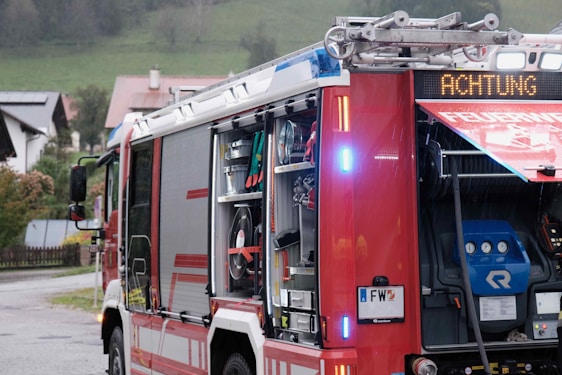 A fire truck is parked on a street with its side compartment open, revealing various firefighting equipment such as hoses, tools, and gauges. A digital sign on the truck displays the word 'ACHTUNG,' indicating a warning or alert. Residential houses and greenery are visible in the background, suggesting a rural area.
