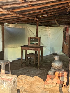 Inside a rustic structure with a thatched roof supported by wooden poles and metal sheeting, there is a small table with an old television on it. The floor appears to be dirt, and there's a stack of bricks with a metallic pot placed on top. A metal stool is visible in the foreground, and the walls are made of white fabric, possibly for insulation or privacy. Posters or paper hang on the wooden wall to the right.