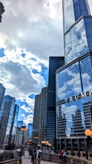 Tall skyscrapers dominate the cityscape under a partly cloudy sky. The glass buildings reflect the surrounding structures and the sky. A prominent building with the text 'TRUMP' is visible in the foreground. Street lights with orange globes line the walkway where people are walking.