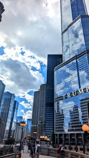 Tall skyscrapers dominate the cityscape under a partly cloudy sky. The glass buildings reflect the surrounding structures and the sky. A prominent building with the text 'TRUMP' is visible in the foreground. Street lights with orange globes line the walkway where people are walking.